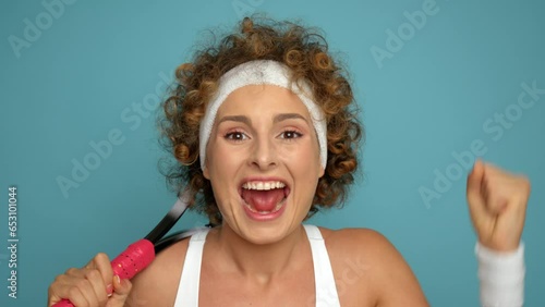 Happy woman with headband on short curly hair celebrates victory in tennis set by raising her fist and waving tennis racket, lifestyle concept, copy space, high quality photo