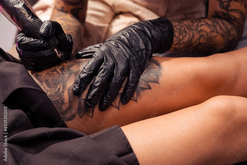 Hands of artist making tattoo of flower design on female thigh