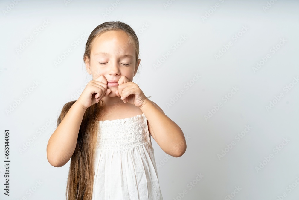 Pleased beautiful kid girl wearing white dress with closed eyes keeps hands near cheeks and smiles tenderly imagines something very pleasant Pleased beautiful kid girl wearing white dress with closed eyes keeps hands near cheeks and smiles tenderly imagines something very pleasant
