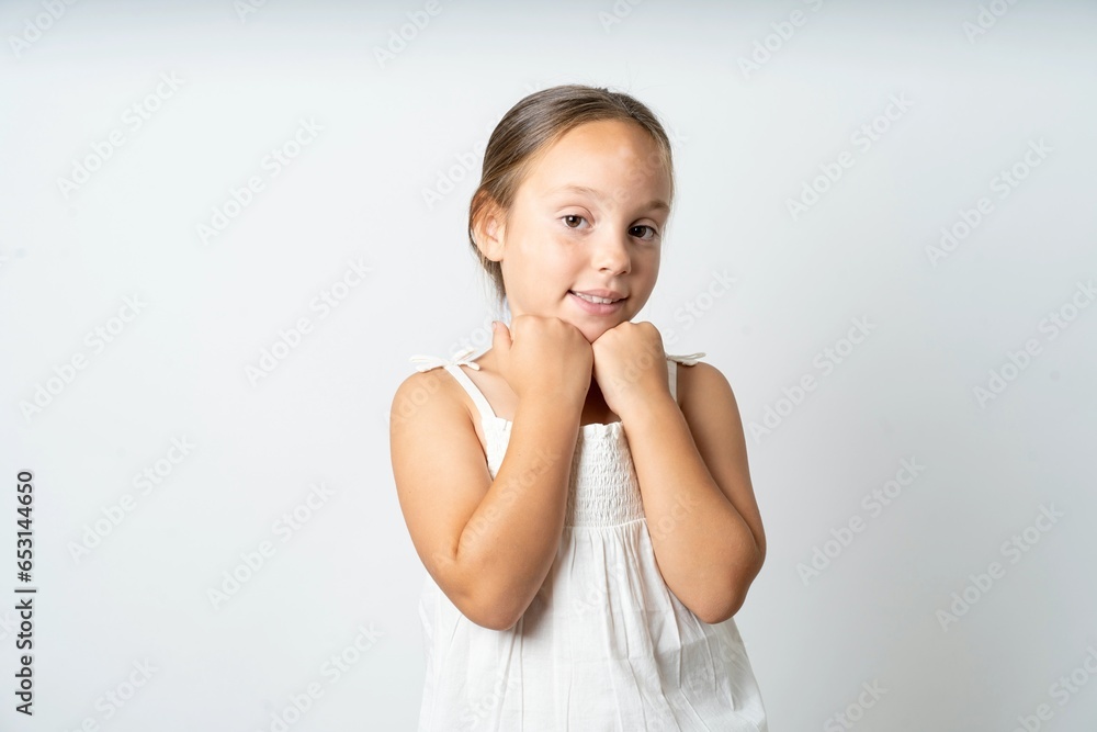 Satisfied beautiful kid girl wearing white dress touches chin with both hands, smiles pleasantly, rejoices good day with lover Satisfied beautiful kid girl wearing white dress touches chin with both hands, smiles pleasantly, rejoices good day with lover