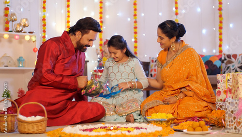 Diwali celebration with gift box exchange - Indian man with his wife and little daughter - Diwali gift  festive background. A happy Indian couple sitting on the floor - Floral rangoli  Diwali rango... photo