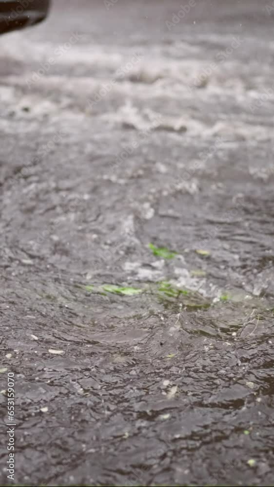 Vertical video, Slow motion, Swift stream of rainwater flows into storm drain, cars are driving at city crossroads on background, panorama. Heavy rain leads to sewage overflow, flood in city