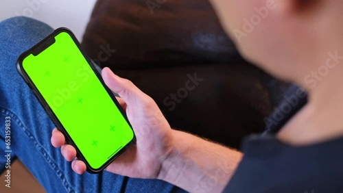 Unrecognizable young man sitting on the couch in a living room and using his smartphone. Scrolling, swiping, touching on a green screen with tracking markers. Browsing on the internet. App template.