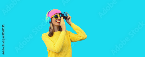 Stylish modern young woman photographer taking a picture with film camera and listens to music in headphones wearing colorful pink hat, yellow sweater on blue studio background