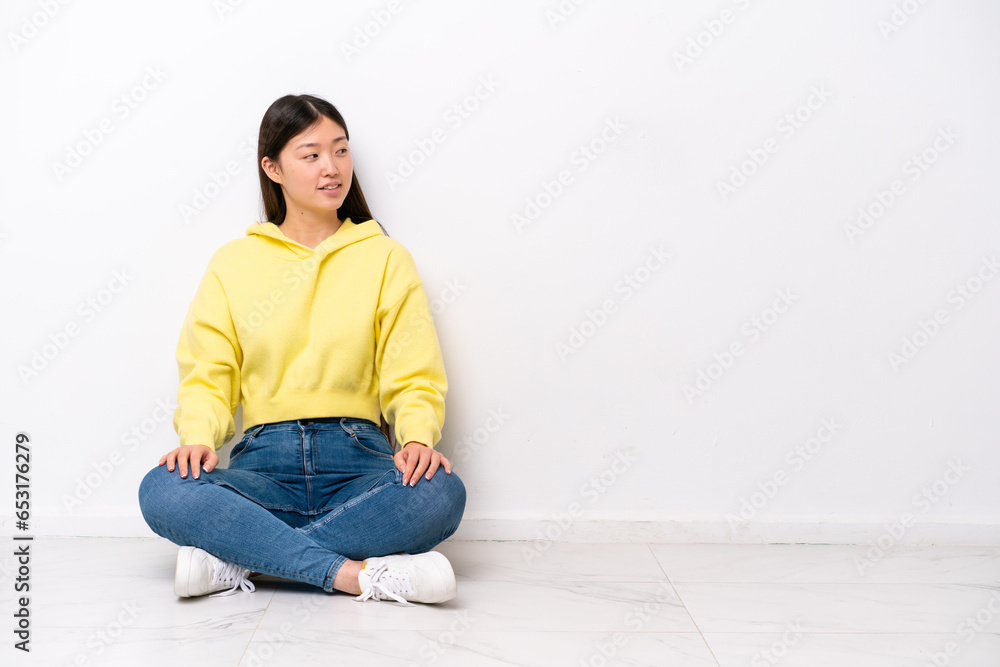 Young Chinese woman sitting on the floor isolated on white wall looking side