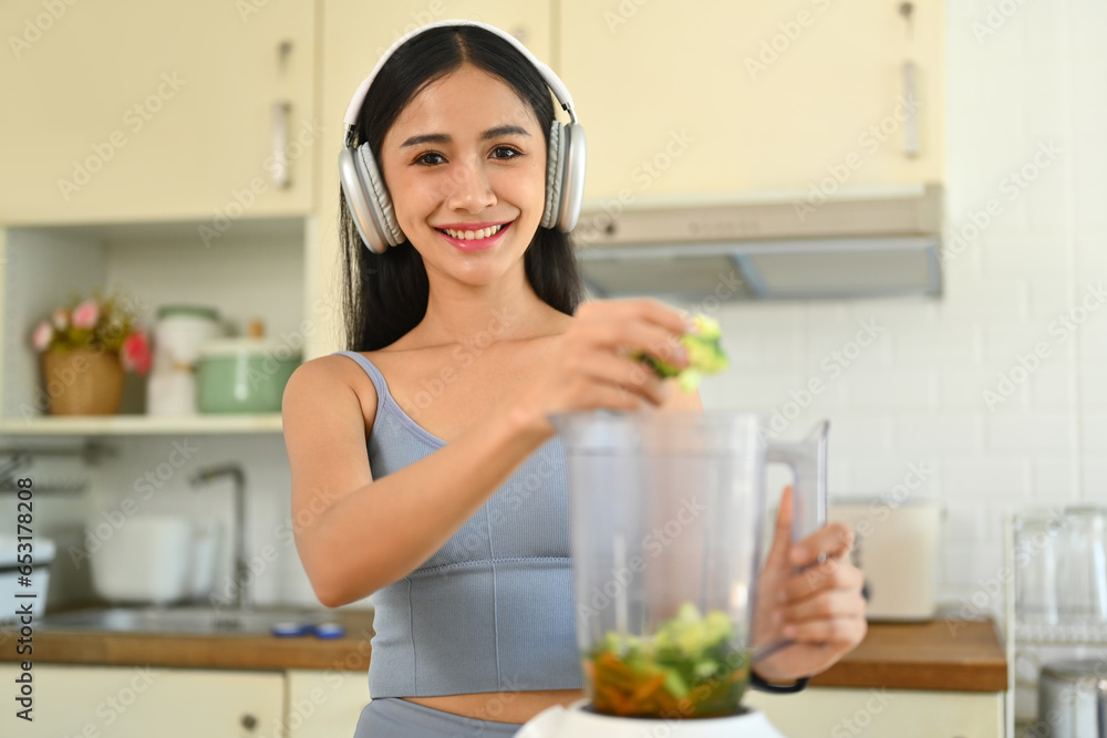 Young woman in fitness clothes making green detox vegetable smoothie with blender in kitchen. Dieting, healthy eating concept