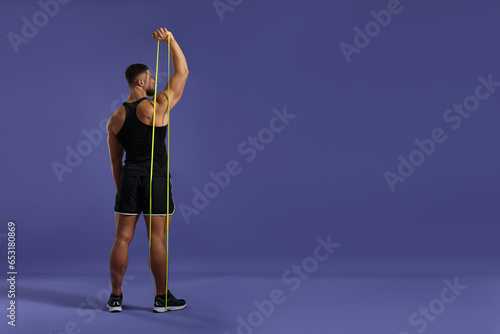 Muscular man exercising with elastic resistance band on purple background, back view. Space for text