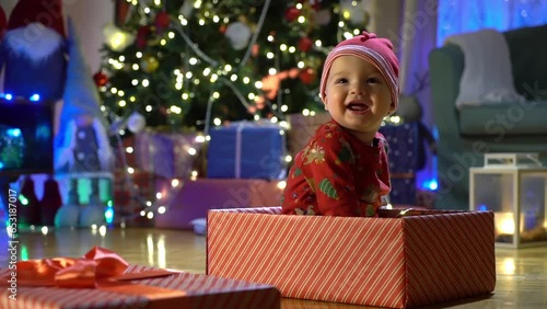 Happy baby boy in a red cap sitting in a gift box on the background of a Christmas tree on Christmas Eve. Happy Holidays, New Year. Cozy warm winter evening at home. Xmas time