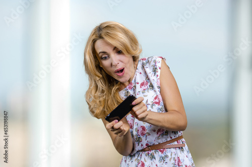 Excited blonde woman plays video game on her smartphone. Blurred window in the background. photo