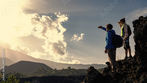 Hiking Couple Admiring Vast Landscape: A mature couple in windbreakers and with backpacks stand atop a rock, taking in the view while shielding their eyes, with an expansive sky perfect for copy. photo