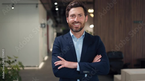Portrait of bearded businessperson crossing arms while posing in lighted office. Handsome caucasian male looking at camera and positively smiling. Concept of facial expression and occupation.