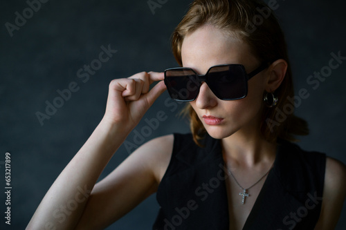 Fashionable beautiful young woman wearing trendy black big butterfly sunglasses, classic vest, posing in studio with dark gray background, natural day light. Copy, empty space for text