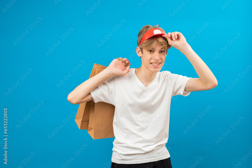 Young seller in casual white t-shirt with paper bag isolated on blue background studio portrait. People lifestyle concept. Mock up copy space. Teen holding package bag with purchases Young seller in casual white t-shirt with paper bag isolated on blue background studio portrait. People lifestyle concept. Mock up copy space. Teen holding package bag with purchases