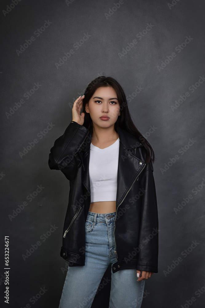 Asian girl in modern clothes stands against gray wall, straightening hair with hand. Brunette with long hair in leather jacket and jeans raised hand up, looking wistfully straight at the camera Asian girl in modern clothes stands against gray wall, straightening hair with hand. Brunette with long hair in leather jacket and jeans raised hand up, looking wistfully straight at the camera