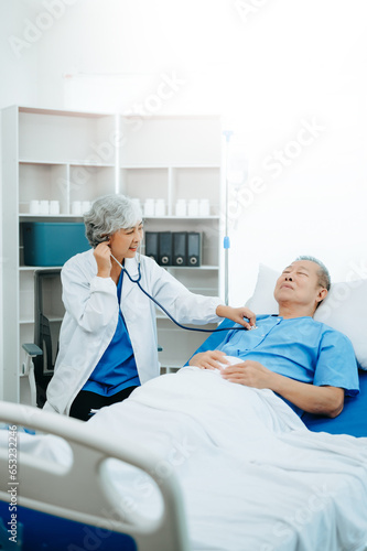 Asian doctor in white suit take notes while discussing and Asian elderly, man patient who lying on bed with receiving saline solution in hospital or clinic.