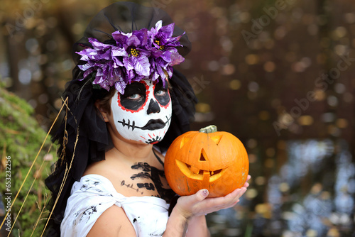 Serious girl in spooky dead bride outfit with pumpkin Jack o lantern looking at camera while standing in nature during Halloween holiday