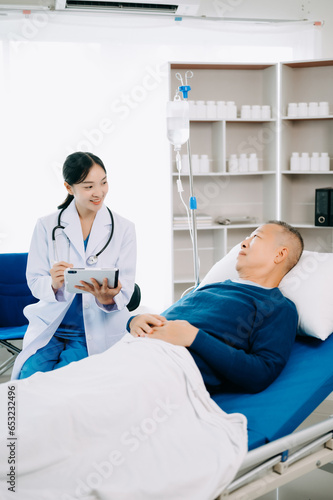 Asian doctor in white suit take notes while discussing and Asian elderly, man patient who lying on bed with receiving saline solution in hospital .