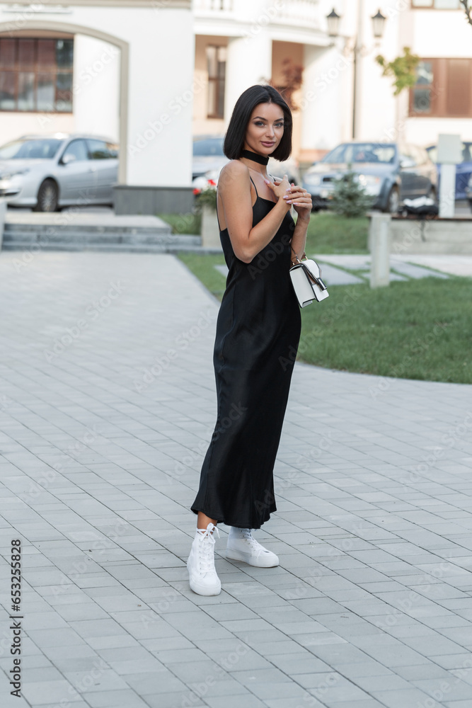 Beautiful elegant fashion woman with a brunette hairstyle in a fashionable long dress with spaghetti straps with a stylish handbag and white sneakers walks in the city. Pretty girl outdoors