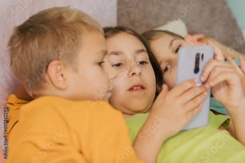 Children play with smartphone at home lying on pillows under the covered blanket