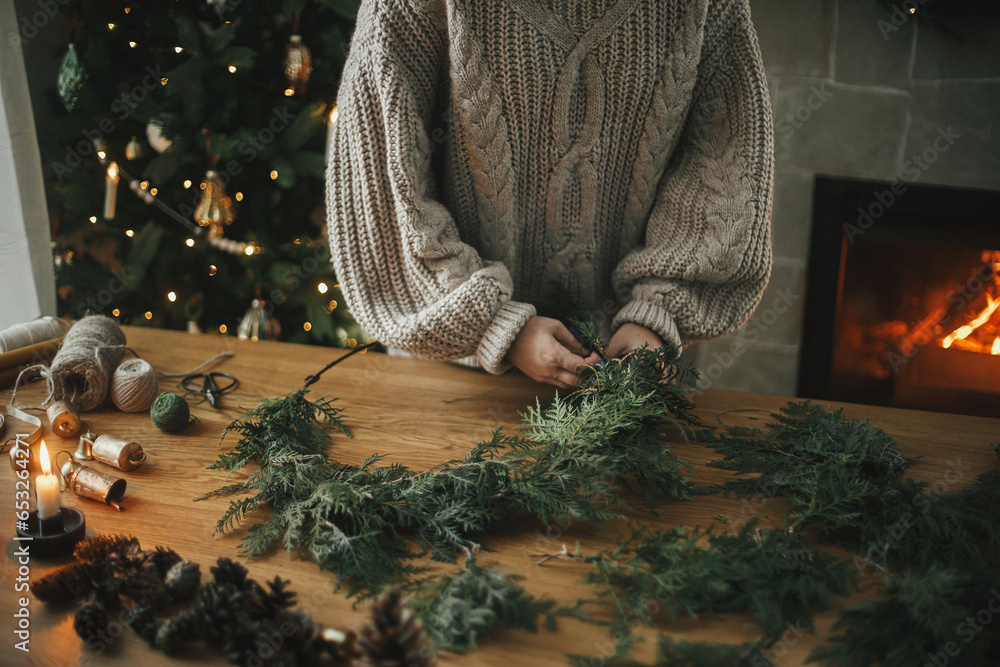 Making Christmas rustic wreath. Hands holding cedar branches, making wreath on wooden table with pine cones, candle, twine, bells in atmospheric festive room. Winter holiday preparations