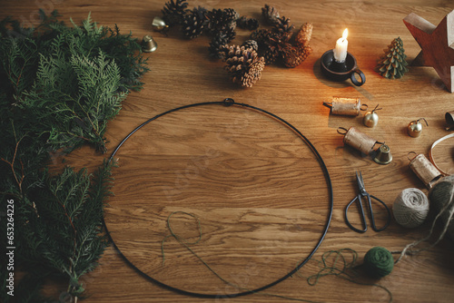Making Christmas rustic wreath. Circle, cedar branches, pine cones, candle, twine, bells on wooden table in moody festive room. Winter holiday preparations, atmospheric time