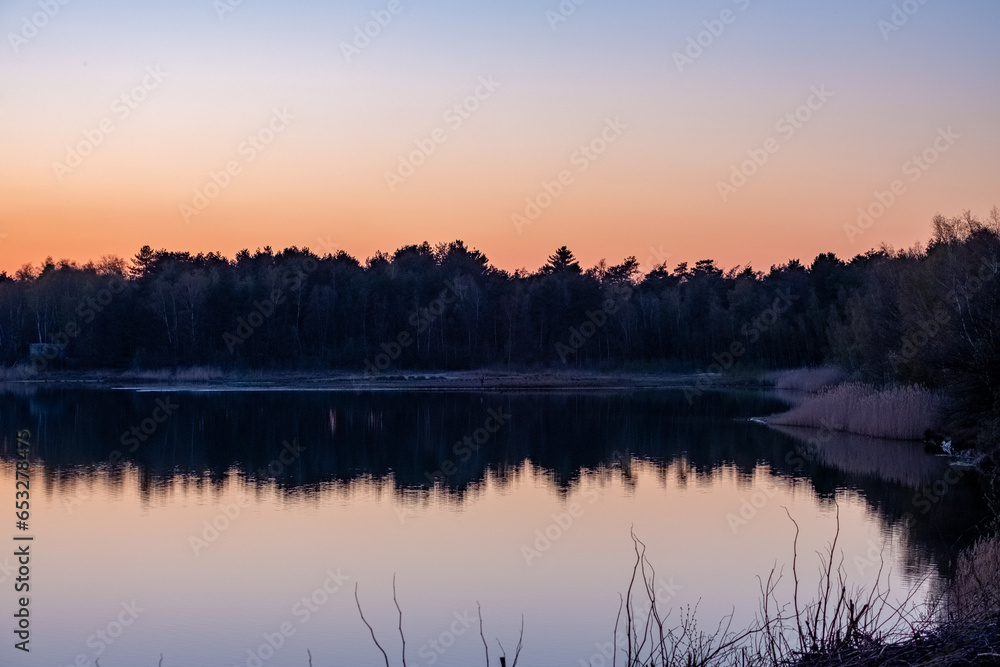 Nature's breathtaking artistry unfolds as a serene forest lake mirrors the dramatic transformation of the sky from vivid blue to fiery orange during dusk. The tranquil waters capture the essence of