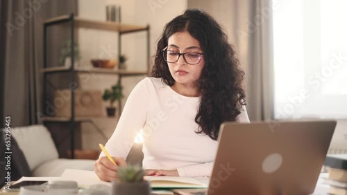 Charming curly girl preparing for exam at university studying at home workplace Pretty student make notes while looking at laptop computer screen indoors Distance remote education