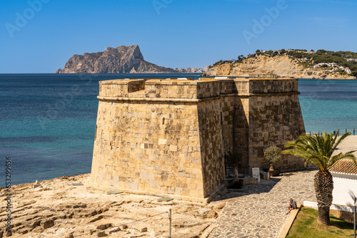 Bild auf Leinwand A panoramic view of the historic stone Moraira Castle on the rocky coast, with the prominent Peñon de Ifach rock formation rising in the distance across the blue sea, Alicante, Spain