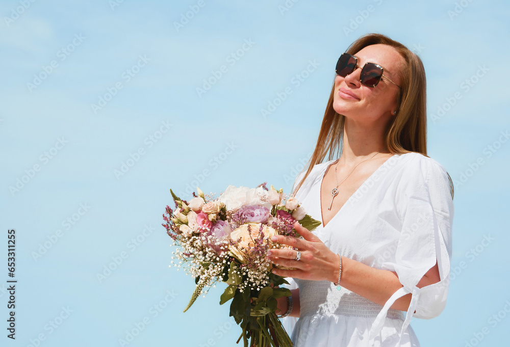 Joyful Young Woman Holding a Bouquet of Flowers on a Beach with Blue Sky Background