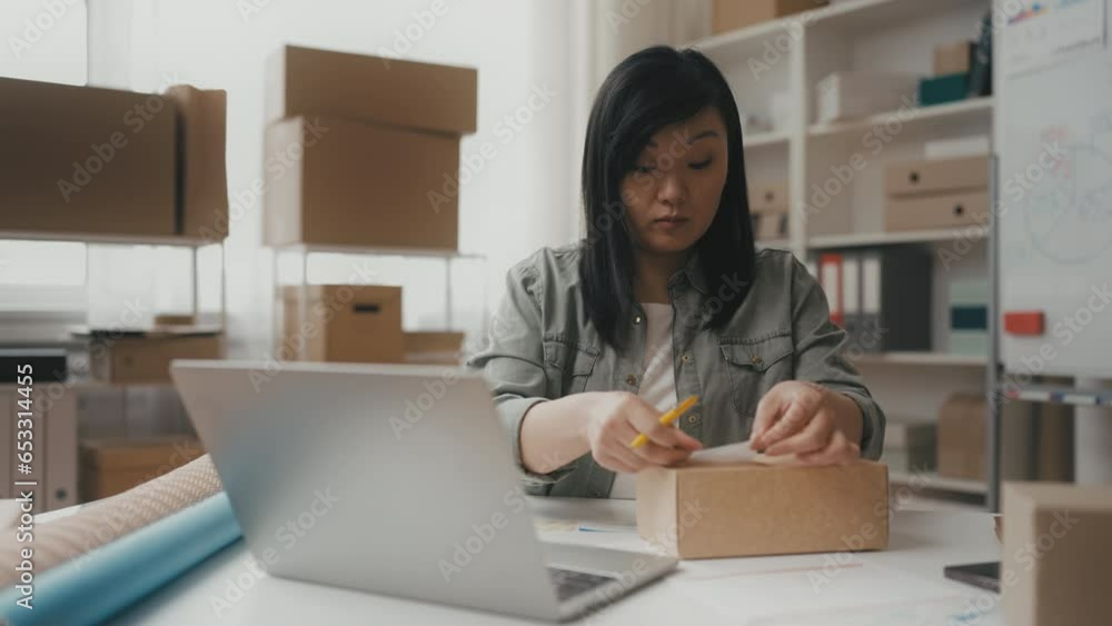Middle-aged Asian woman entrepreneur putting sticker on box, packing orders Middle-aged Asian woman entrepreneur putting sticker on box, packing orders
