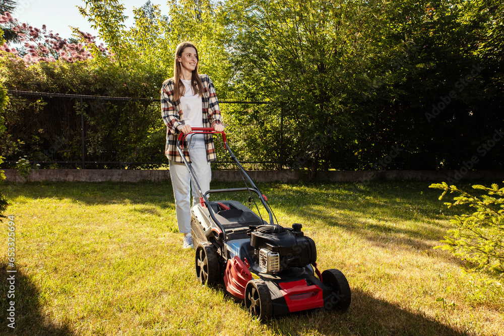 Fototapeta premium Female gardener working in autumn, cutting grass in backyard. Concept of gardening, work, nature. Housework, gardening and country life. Home garden grass cutting woman mowing with lawn mower.