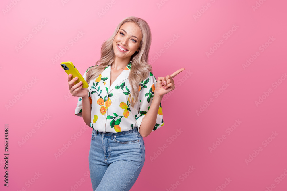 Portrait of positive woman with curly hairdo wear stylish shirt hold smartphone indicating empty space isolated on pink color background Portrait of positive woman with curly hairdo wear stylish shirt hold smartphone indicating empty space isolated on pink color background