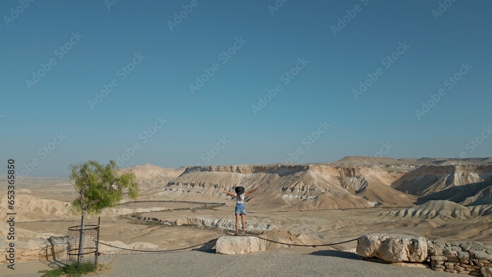 European woman stands against the background of the Negev Desert in Israel is visible in the background over mountains, sandy hills and rock formations.