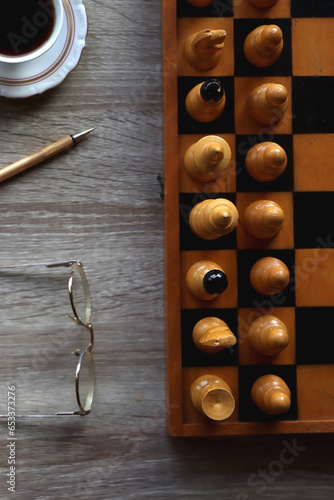 Vintage wooden chessboard, books, glasses, pen, cup of tea or coffee and scented candle on the table. Dark academia concept. Top view.