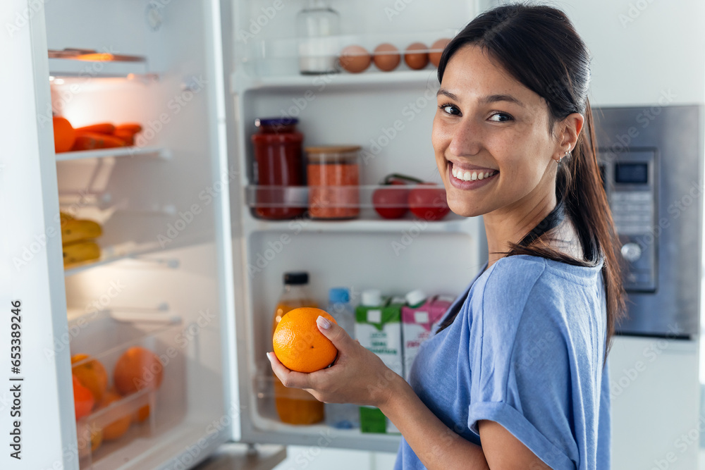 Beautiful young woman taking an orange from the fridge while looking at camera in the kitchen at home Beautiful young woman taking an orange from the fridge while looking at camera in the kitchen at home