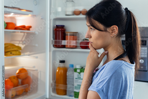 Shot of pretty young woman hesitant to eat in front of the fridge in the kitchen. photo