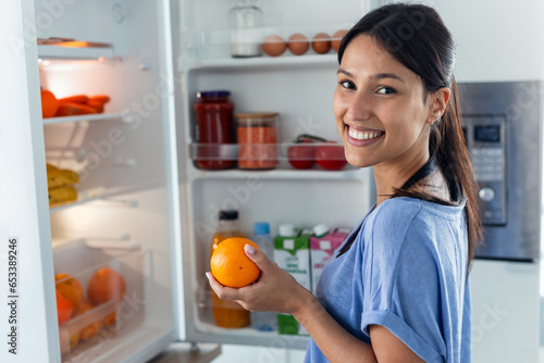 Beautiful young woman taking an orange from the fridge while looking at camera in the kitchen at home