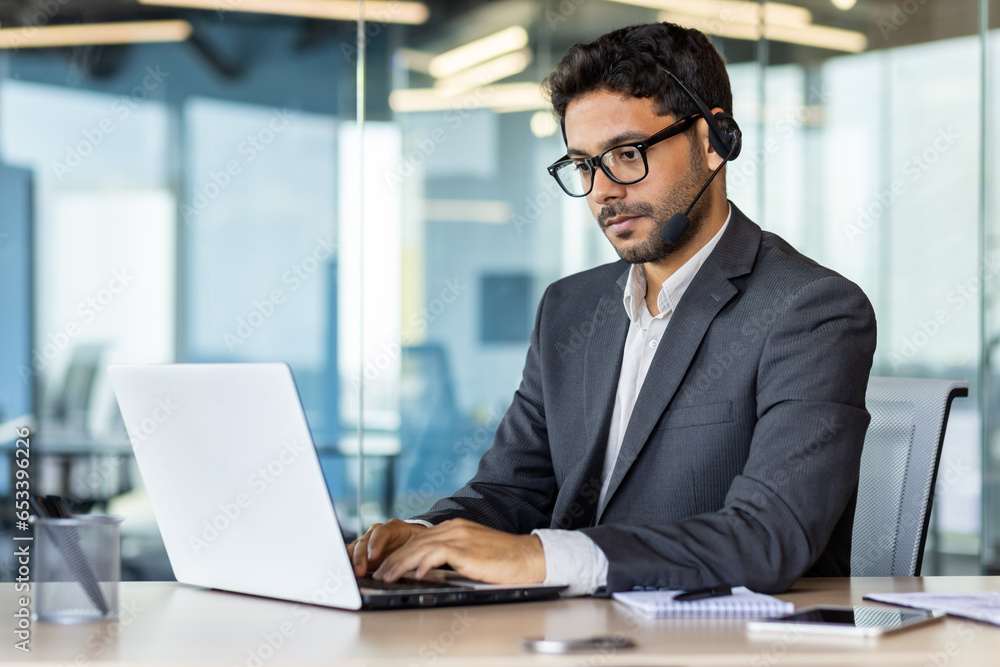 A young Indian businessman is working in the office on a laptop and wearing headphones. Concentrated typing on the keyboard, chats. A young Indian businessman is working in the office on a laptop and wearing headphones. Concentrated typing on the keyboard, chats.