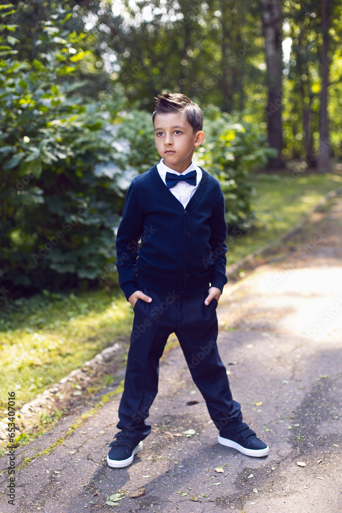 first-grader boy in a uniform with a bow tie is standing on the street with greenery. the child goes to school first-grader boy in a uniform with a bow tie is standing on the street with greenery. the child goes to school