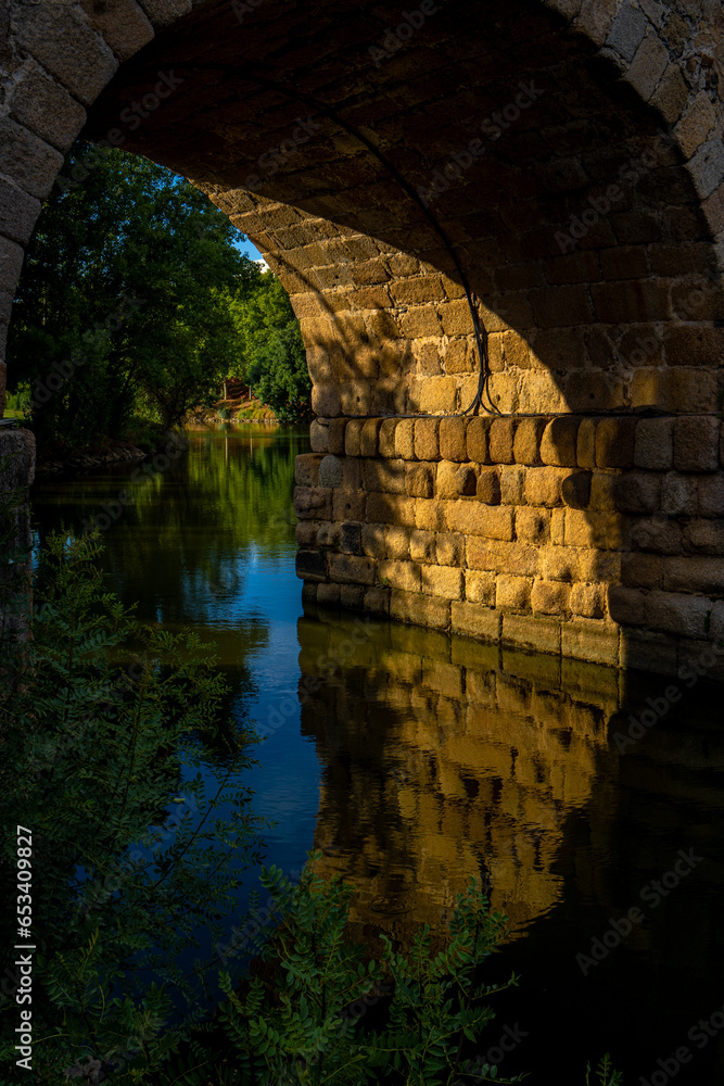 Arch partially illuminated by evening sunlight of Mérida's Roman bridge over the river, with its reflection in the water.