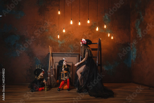 Girls in witch costumes are sitting near the fireplace making a potion, and their mother is sitting nearby.