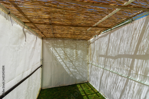 A white building of a public Sukkah, before the Sukkot holiday, in Ein Hamad Park in Jerusalem. The Sukkah is made of wood and branches, and it is covered with a roof of leaves.