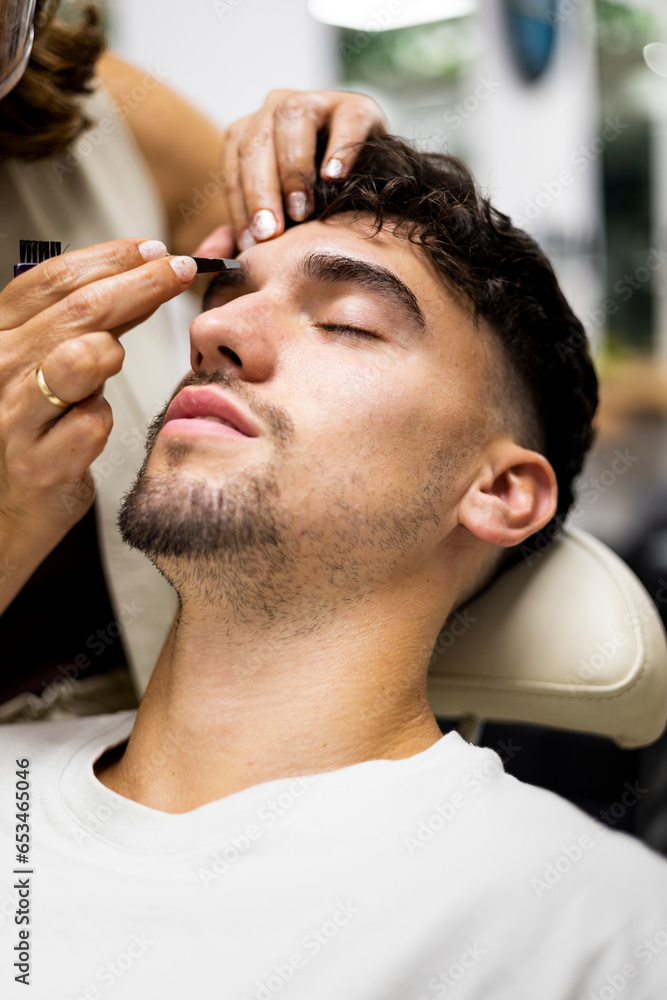 Obraz premium Vertical photo of a bearded young adult with his eyes closed is sitting in a beauty center cutting his eyebrows.The woman stylist uses tweezers to pull hairs from the eyebrows.Removal of eyebrow hairs