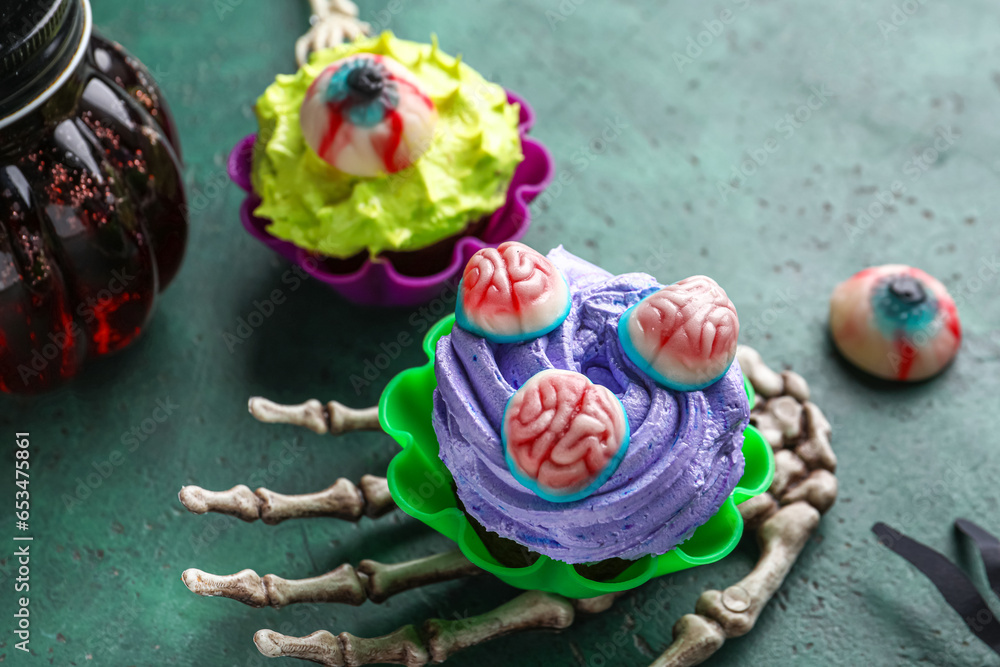 Skeleton hand with delicious Halloween cupcakes and candies on green background, closeup