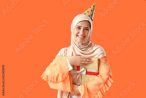 Young Muslim woman in party hat with gift on orange background