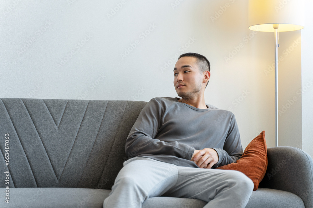Portrait of young, pensive Asian man sitting on sofa in apartment in the living room, looking away