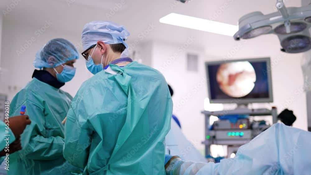 Rear view of surgeons in the operational room. Nurse passes the tool to a doctor. Screen works at the backdrop. Rear view of surgeons in the operational room. Nurse passes the tool to a doctor. Screen works at the backdrop.