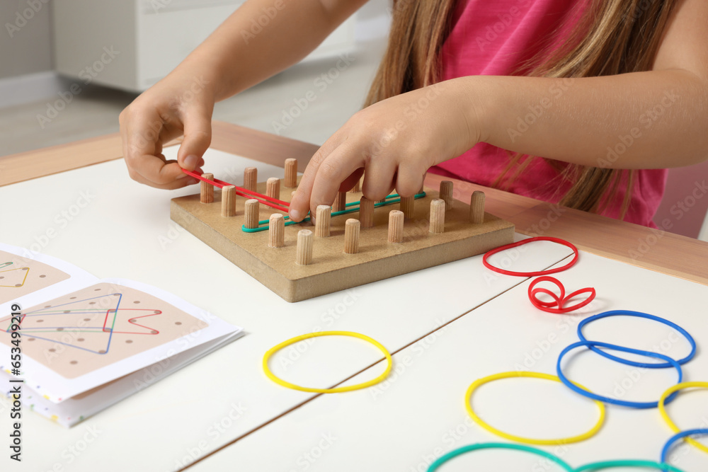 Fototapeta premium Motor skills development. Girl playing with geoboard and rubber bands at white table indoors, closeup