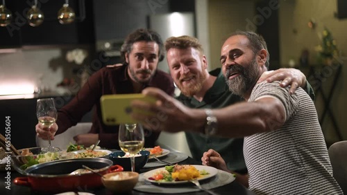 Group of middle aged men taking a selfie on a smartphone while having lunch and wine together in the kitchen at home