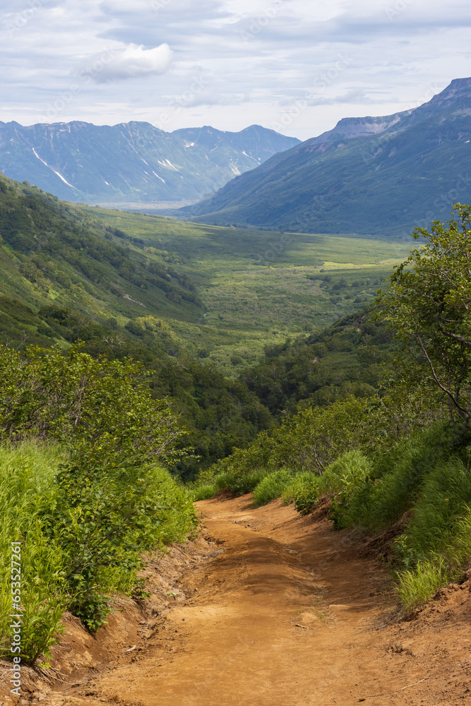 Summer mountain landscape. Dirt road on a mountain slope. View from the hiking trail to the mountain valley. Travel and hiking on the Kamchatka Peninsula. Kamchatka Territory, Far East of Russia.